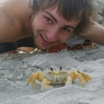 A smiling man crouches down to pose on a sandy beach with a ghost crab